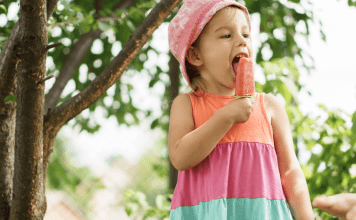 A little girl in a pink hat, eating a mango smoothie popscicle.