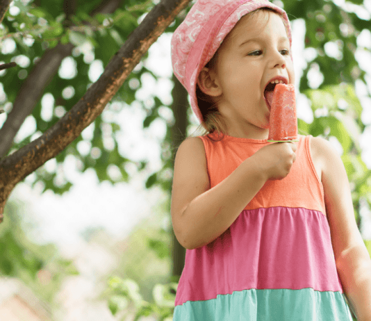 A little girl in a pink hat, eating a mango smoothie popscicle.