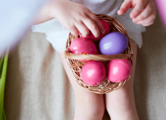A child sitting down with a basket of pink and purple plastic eggs in their lap.