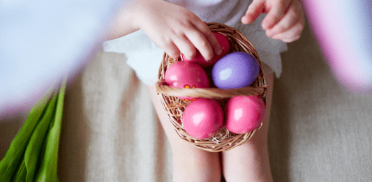 A child sitting down with a basket of pink and purple plastic eggs in their lap.