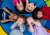 Six young children laying on the ground on a colorful rug