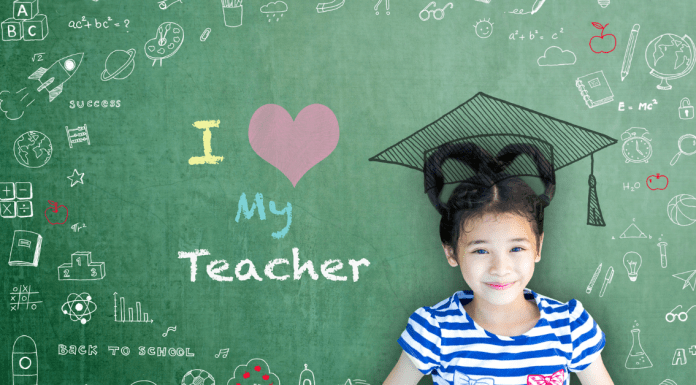 A little girl, smiling in front of a chalkboard that says 'I love my teacher'