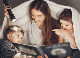 A mom reading a book to her two sons. They are smiling, underneath a sheet, using a flashlight to read.