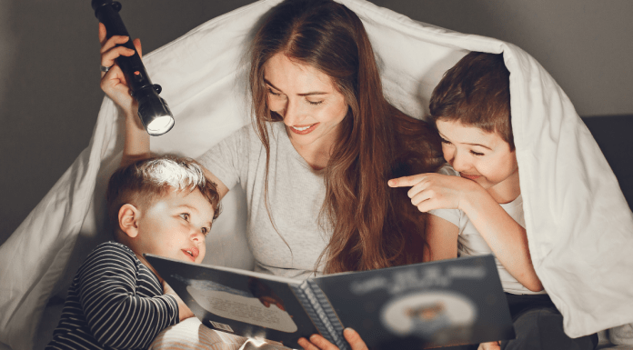 A mom reading a book to her two sons. They are smiling, underneath a sheet, using a flashlight to read.