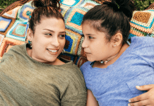 A mom and her daughter laying on a blanket talking and smiling.