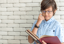 A young boy with glasses reading a book with a smile on his face.